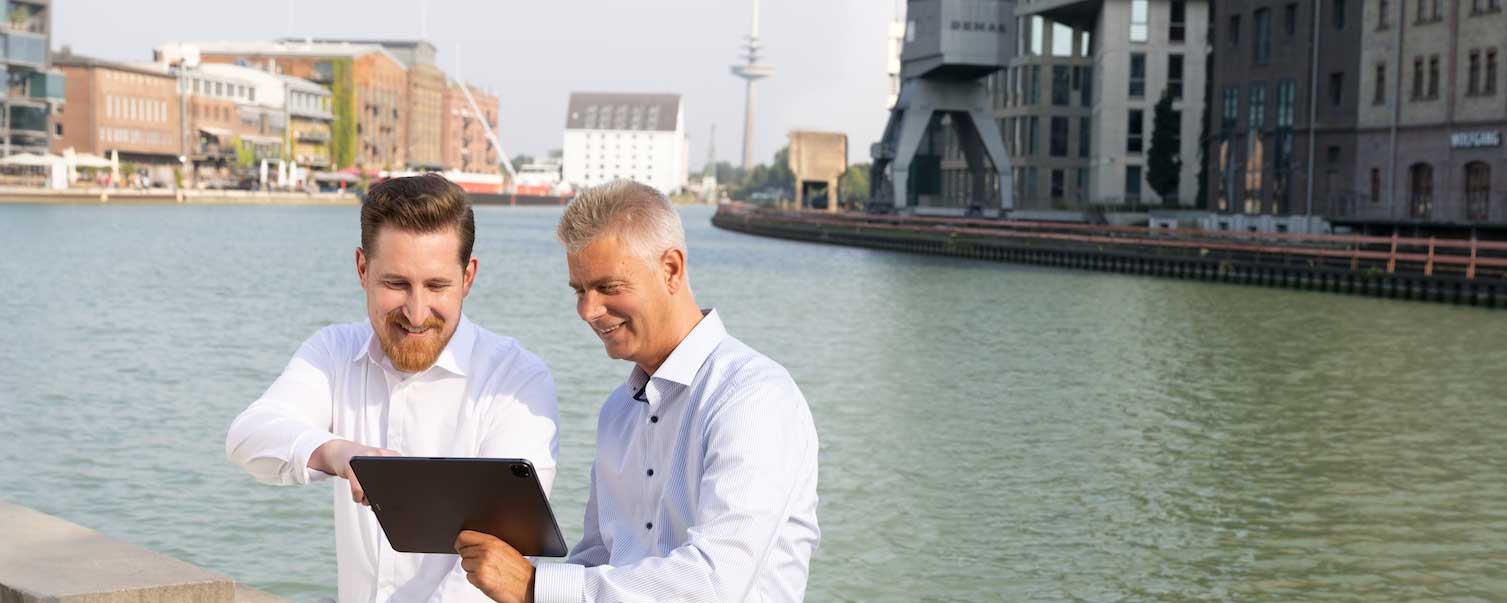 Zwei Männer schauen auf ein Tablett und sitzen am Ufer des Stadthafens Münster